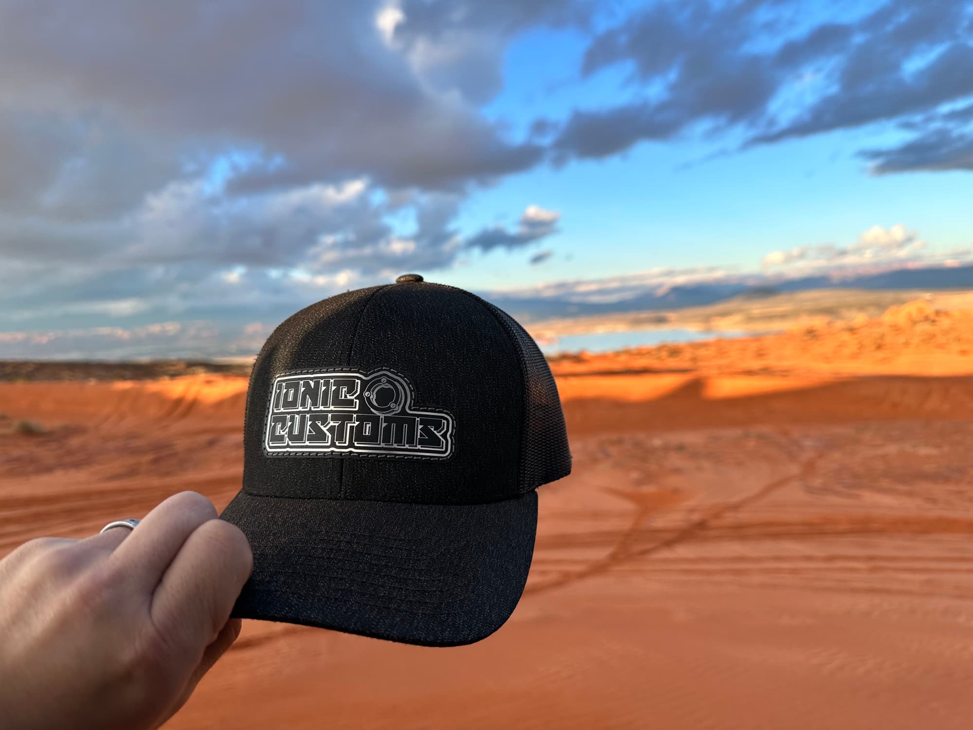 Ionic Customs leather patch trucker hat held up against desert dunes at sunset